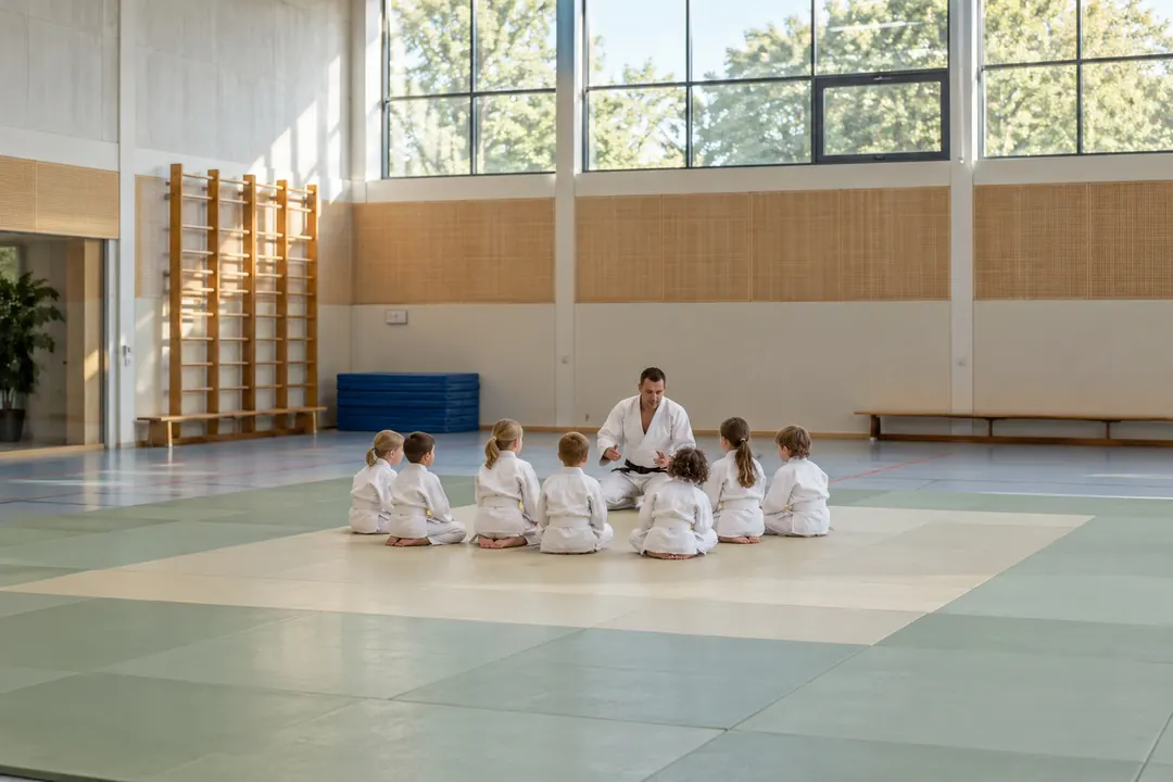 Een judoleraar zit in de zaal op de mat tegenover een groep kinderen in judopak